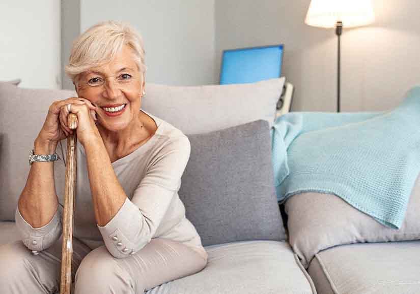 Woman in beige clothes on beige couch leaning on cane smiling
